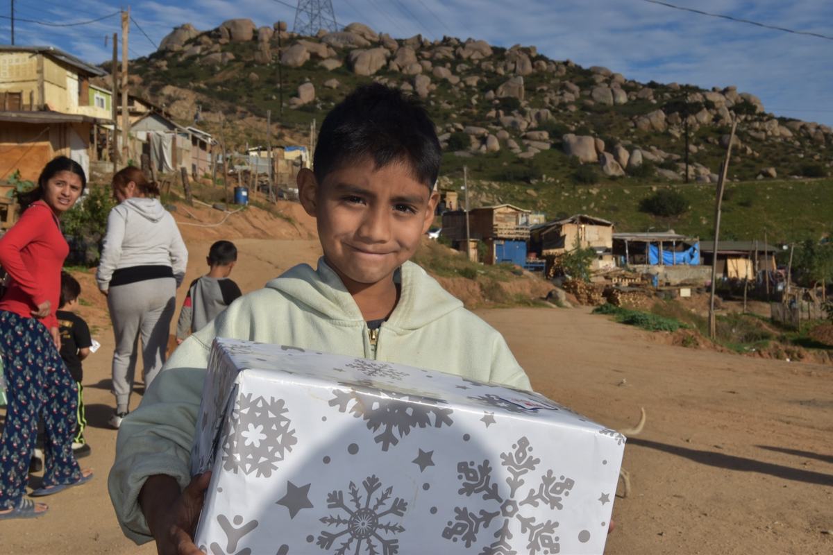 A boy holding a wrapped gift with his village behind him
